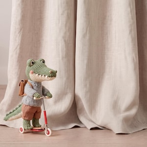 Könnte beinhalten: Ein gehäkeltes grünes Krokodil-Stofftier, das einen grauen Strickpullover und eine braune Shorts trägt sowie einen kleinen beigen Rucksack aufhat. Das Krokodil steht auf einem rot-silbernen Spielzeugroller mit weißen Rädern und Garnstruktur.