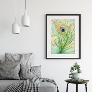 May include: A watercolor painting of a peacock feather with a blue eye, surrounded by green and yellow lines and water droplets. The painting is framed in black and hanging on a white wall.