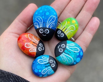 May include: A collection of hand-painted rocks shaped like colorful bugs. Each rock features a different color scheme, including blue, red, green, and turquoise, with white and black details. The bugs have smiling faces and decorative wing patterns.