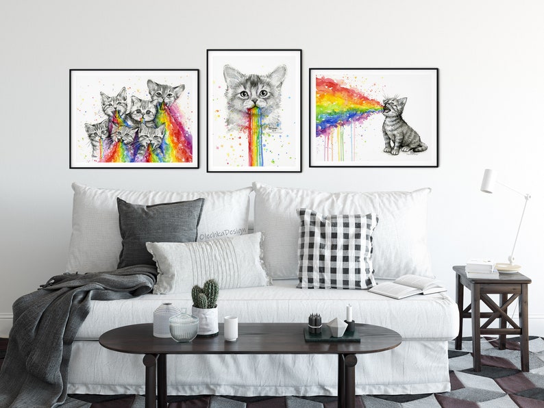 May include: Three framed watercolor paintings of cats vomiting rainbows. The paintings are hung on a white wall above a white couch with gray throw pillows and a coffee table.