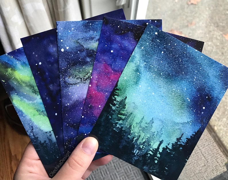 May include: Five watercolor paintings on cardstock featuring different starry night skies. The paintings are all in shades of blue, green, and purple, with white stars scattered across the canvas. Some of the paintings feature a silhouette of a forest in the foreground.