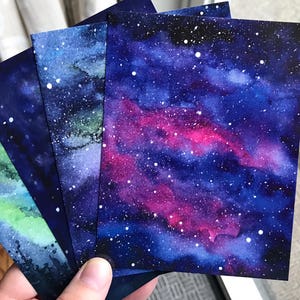 May include: Four watercolor paintings of a galaxy with white stars on a dark blue background. One painting has a pink and purple nebula, one has a green and blue nebula, and two have a dark blue nebula.