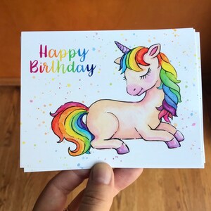 Pode incluir: Um cart&atilde;o branco com uma ilustra&ccedil;&atilde;o de um unic&oacute;rnio arco-&iacute;ris e o texto "Happy Birthday" em letras coloridas. O unic&oacute;rnio tem uma crina e uma cauda arco-&iacute;ris e est&aacute; deitado com os olhos fechados.