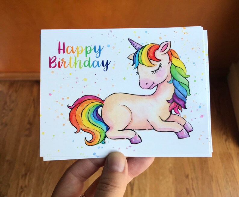 Pode incluir: Um cart&atilde;o branco com uma ilustra&ccedil;&atilde;o de um unic&oacute;rnio arco-&iacute;ris e o texto "Happy Birthday" em letras coloridas. O unic&oacute;rnio tem uma crina e uma cauda arco-&iacute;ris e est&aacute; deitado com os olhos fechados.