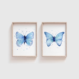 May include: Two framed watercolor paintings of blue butterflies on a white background. Each butterfly is surrounded by small blue and green dots.