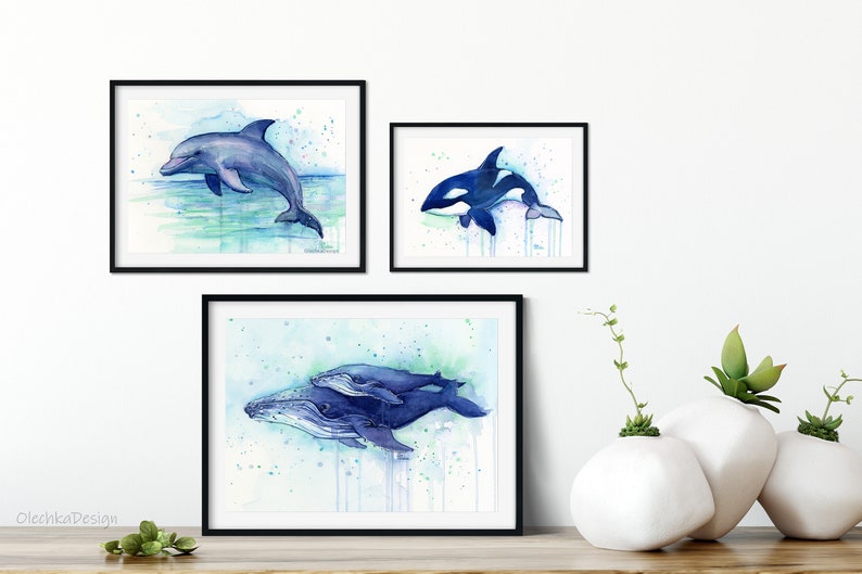 May include: Three watercolor paintings of marine animals in black frames. The top left painting shows a dolphin jumping out of the water. The top right painting shows an orca swimming. The bottom painting shows a humpback whale and calf swimming.