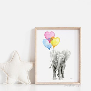 May include: A framed watercolor print of a gray elephant holding heart-shaped balloons in pink, blue, and yellow. The artwork is in a light wood frame and is set against a white background. A white star-shaped pillow is to the left.