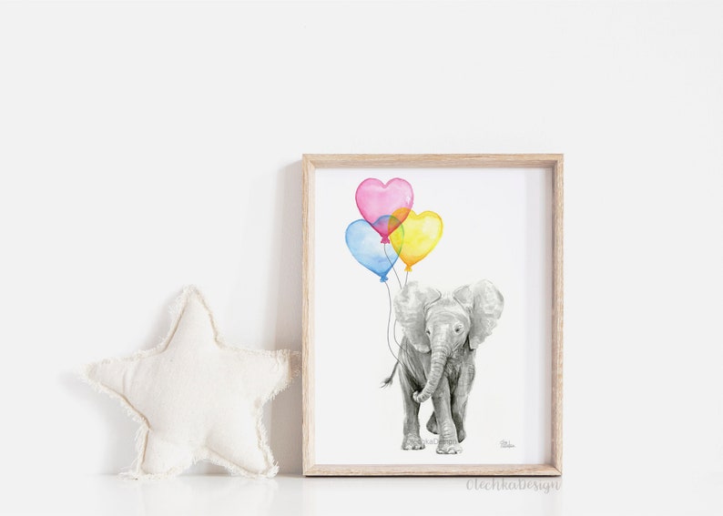 May include: A framed watercolor print of a gray elephant holding heart-shaped balloons in pink, blue, and yellow. The artwork is in a light wood frame and is set against a white background. A white star-shaped pillow is to the left.