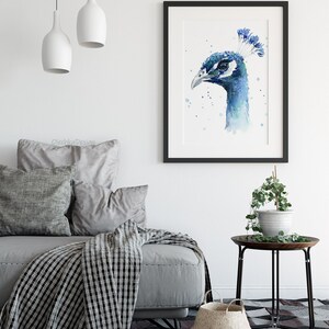 May include: A watercolor painting of a peacock's head in shades of blue and green, framed in black and displayed on a white wall above a gray sofa with pillows and a black and white throw blanket.