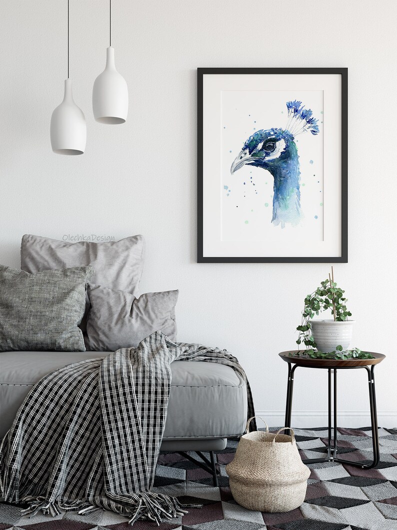 May include: A watercolor painting of a peacock's head in shades of blue and green, framed in black and displayed on a white wall above a gray sofa with pillows and a black and white throw blanket.