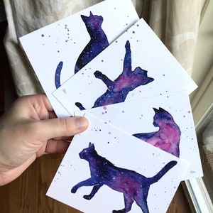 May include: Four watercolor paintings of cats with a galaxy design. The cats are in different poses, including sitting, lying down, and jumping. The galaxy design is in shades of purple, blue, and pink.