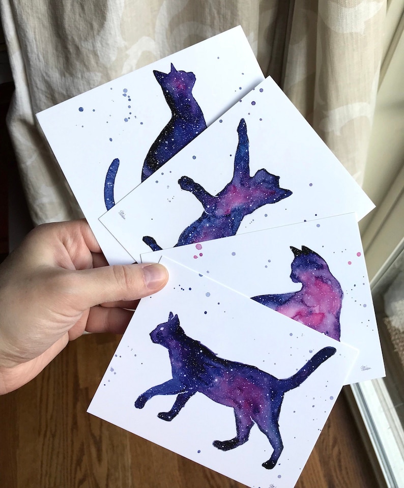 May include: Four watercolor paintings of cats with a galaxy design. The cats are in different poses, including sitting, lying down, and jumping. The galaxy design is in shades of purple, blue, and pink.
