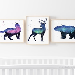 May include: Three watercolor prints featuring silhouettes of a cat, deer, and bear with a starry night sky and forest scene inside each animal. The prints are framed in natural wood.