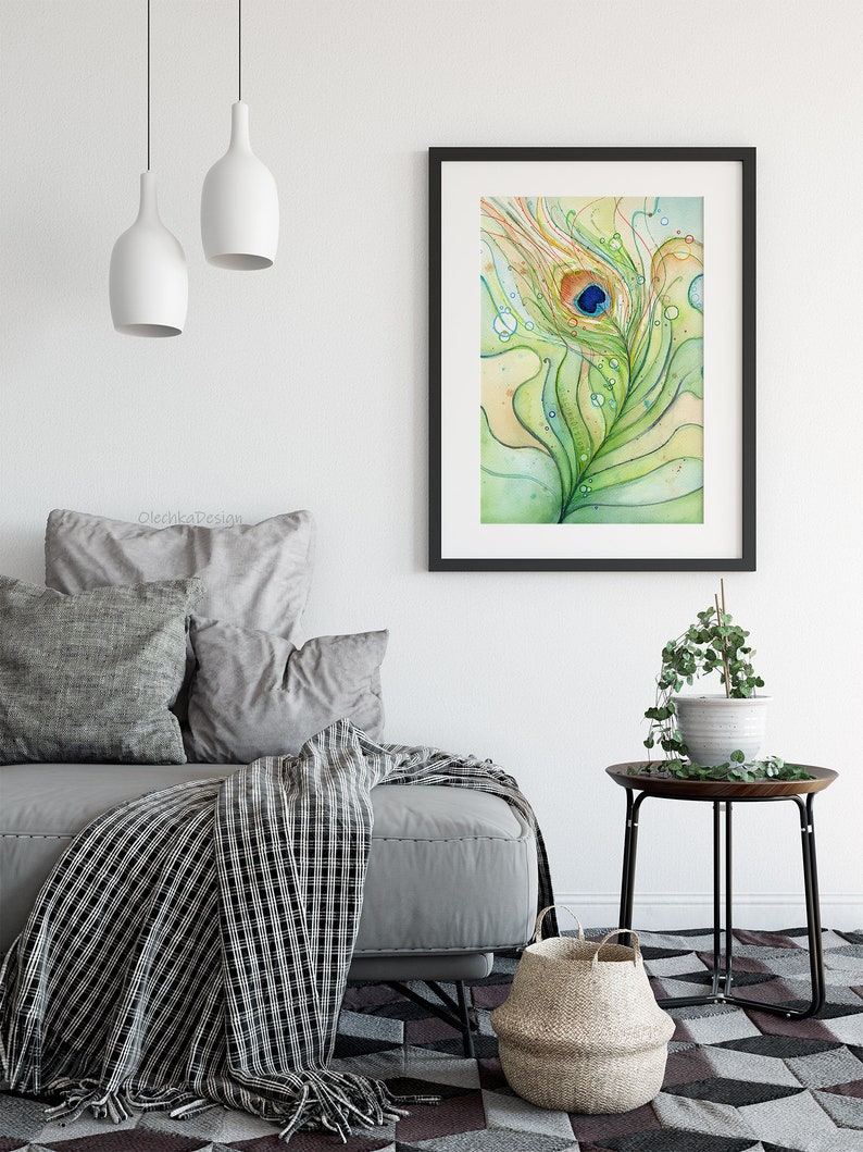 May include: A watercolor painting of a peacock feather with a blue eye, surrounded by green and yellow swirls and water droplets. The painting is framed in black and hanging on a white wall above a gray couch.