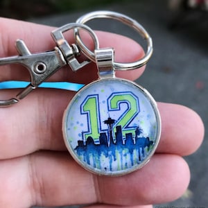 May include: A silver keychain with a round glass charm featuring a green and blue graphic of the number 12, a skyline, and the Space Needle. The charm is attached to a silver split ring and a silver lobster claw clasp.