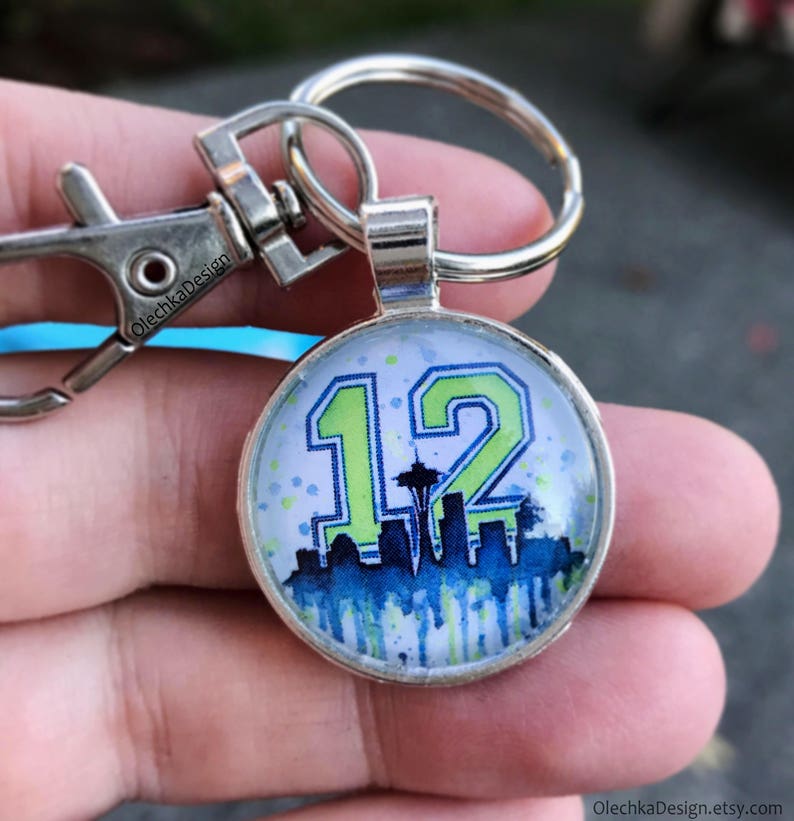 May include: A silver keychain with a round glass charm featuring a green and blue graphic of the number 12, a skyline, and the Space Needle. The charm is attached to a silver split ring and a silver lobster claw clasp.