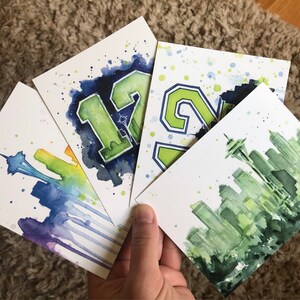 May include: Four watercolor paintings on white cardstock. The paintings depict a Seattle skyline, the number 12, the number 24, and a Seattle skyline with a rainbow.
