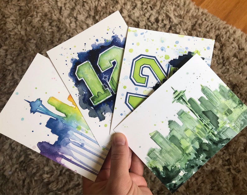 May include: Four watercolor paintings on white cardstock. The paintings depict a Seattle skyline, the number 12, the number 24, and a Seattle skyline with a rainbow.