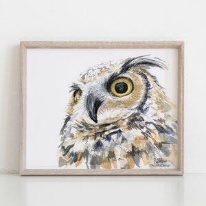 May include: Watercolor painting of an owl's face. The owl has large, yellow eyes and brown and gray feathers. The painting is framed in a light wood frame.