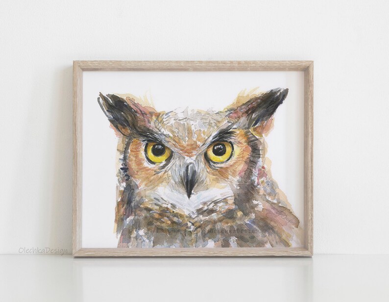 May include: A watercolor painting of an owl's face. The owl has large, yellow eyes and brown and gray feathers. The painting is framed in a light wood frame.