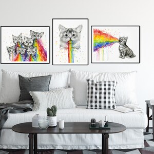 May include: Three framed watercolor paintings of cats vomiting rainbows. The paintings are hung on a white wall above a white couch with gray throw pillows and a coffee table.