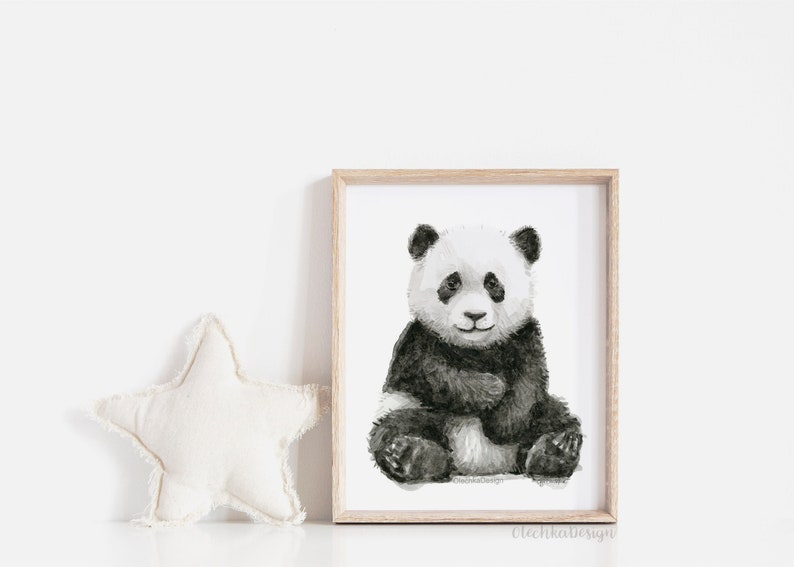 May include: A framed watercolor print of a panda bear sitting. The panda is black and white with a soft, textured appearance. The frame is light-colored wood. A white star-shaped pillow is to the left of the frame.