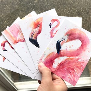 May include: Set of six watercolor paintings of pink flamingos. Each painting features a different pose of the flamingo, with some showing the head and neck, while others show the entire bird. The paintings are done in a loose, impressionistic style, with splashes of color and white space.