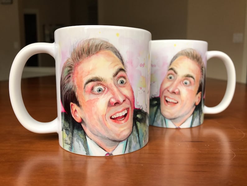 May include: Two white ceramic mugs with a watercolor illustration of a man's face. The man has a surprised expression with his mouth open and his eyes wide. The background is a light pink with yellow and white watercolor splatters.