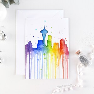 May include: A watercolor painting of the Seattle skyline in a rainbow of colors. The Space Needle is the focal point, with colorful drips of paint flowing down. The artwork is on a white card with the text "OlechkaDesign".