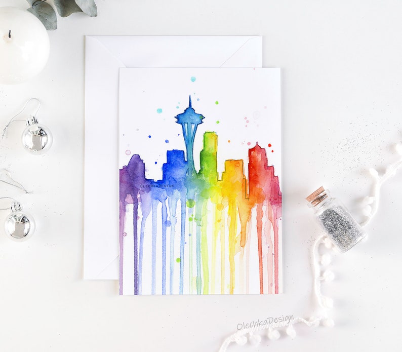 May include: A watercolor painting of the Seattle skyline in a rainbow of colors. The Space Needle is the focal point, with colorful drips of paint flowing down. The artwork is on a white card with the text "OlechkaDesign".