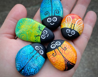 May include: Five painted rocks with ladybug designs. The ladybugs are painted in various colors, including blue, green, yellow, orange, and black. Each ladybug has a unique design and features black eyes and a smiling face.
