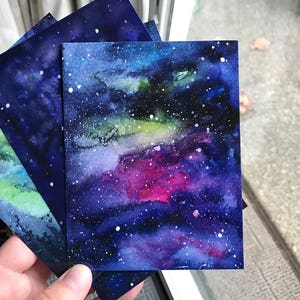 May include: Four watercolor paintings of a galaxy with a dark blue background and white stars. The galaxy has shades of green, pink, and purple.