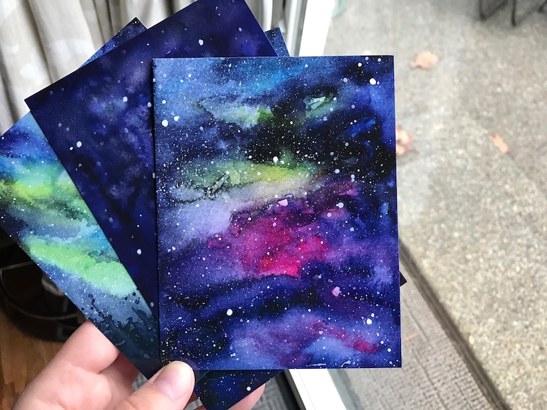 May include: Four watercolor paintings of a galaxy with a dark blue background and white stars. The galaxy has shades of green, pink, and purple.