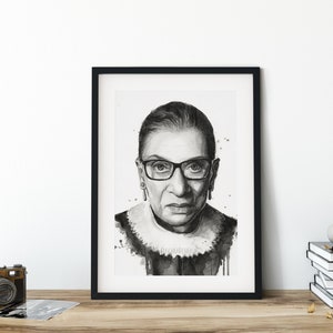 May include: Black and white framed print of a portrait of Ruth Bader Ginsburg. The print is in a black frame with a white mat. The portrait is a close-up of Ginsburg's face, and she is wearing glasses.