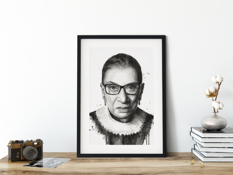 May include: Black and white framed print of a portrait of Ruth Bader Ginsburg. The print is in a black frame with a white mat. The portrait is a close-up of Ginsburg's face, and she is wearing glasses.