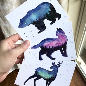 May include: Three watercolor prints featuring silhouettes of a bear, a cat, and a deer. Each silhouette is filled with a galaxy-like watercolor design in shades of blue, purple, and green.