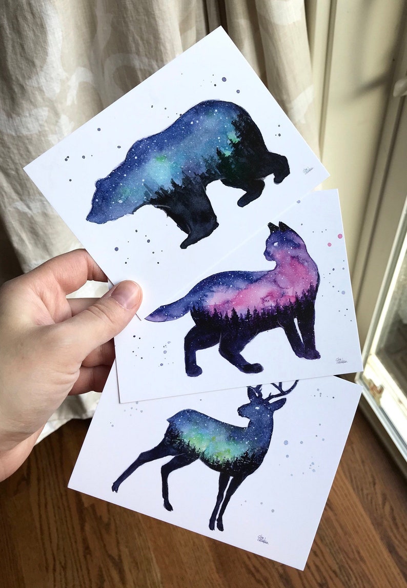 May include: Three watercolor prints featuring silhouettes of a bear, a cat, and a deer. Each silhouette is filled with a galaxy-like watercolor design in shades of blue, purple, and green.