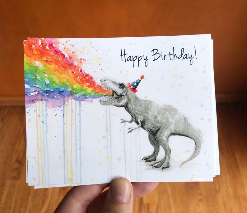 May include: A watercolor illustration of a dinosaur wearing a party hat and blowing a rainbow out of its mouth. The text "Happy Birthday!" is written in black at the top of the card.