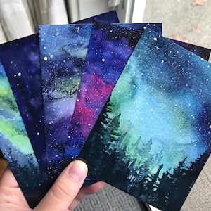 May include: Five watercolor paintings on paper, each depicting a different night sky scene. The scenes include a green and blue aurora borealis, a purple and blue galaxy, a pink and blue galaxy, and two different blue and green starry skies with a silhouette of a forest in the foreground.