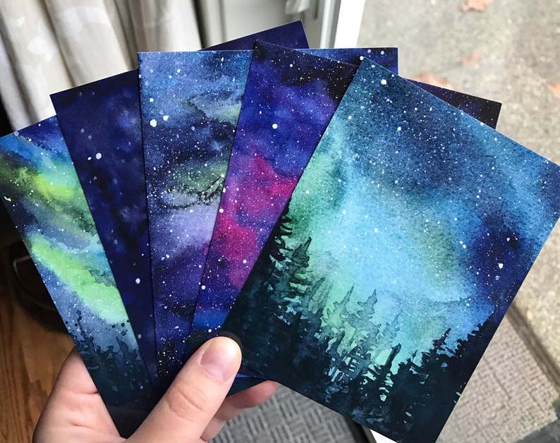 May include: Five watercolor paintings on paper, each depicting a different night sky scene. The scenes include a green and blue aurora borealis, a purple and blue galaxy, a pink and blue galaxy, and two different blue and green starry skies with a silhouette of a forest in the foreground.