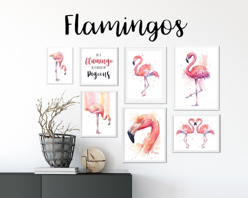 May include: A set of 7 framed watercolor paintings featuring pink flamingos. The paintings are in white frames and are arranged on a white wall. One of the paintings has the text "Be a flamingo in a flock of pigeons".