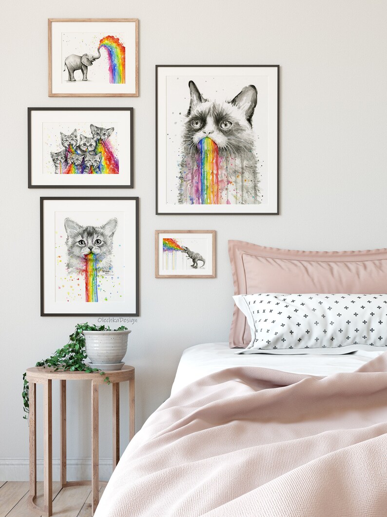 May include: A bedroom interior with a pink bedspread and a white pillow with black crosses. There are five framed black and white watercolor paintings on the wall, featuring a cat, an elephant, and kittens, all vomiting rainbows.