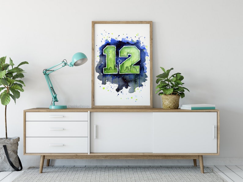 May include: A framed watercolor print featuring the number "12" in green, set against a blue and green watercolor background. The artwork is displayed on a white cabinet with a wooden frame, complemented by a teal desk lamp and potted plants.