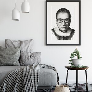 May include: Black and white portrait print of Ruth Bader Ginsburg in a black frame. The print is on a white background and features a watercolor effect. The print is hanging on a white wall above a gray couch with pillows and a black and white throw blanket.