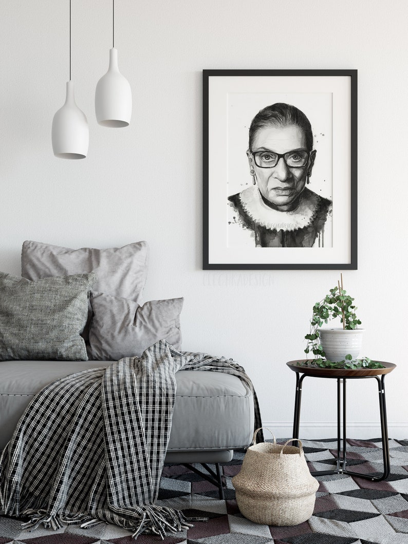 May include: Black and white portrait print of Ruth Bader Ginsburg in a black frame. The print is on a white background and features a watercolor effect. The print is hanging on a white wall above a gray couch with pillows and a black and white throw blanket.