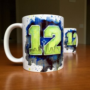 May include: Two white ceramic mugs with a green and yellow "12" design, a cityscape, and a blue and gray watercolor background. The mugs are on a wooden surface.