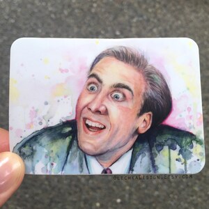 Op de afbeelding: Een aquarel schilderij van Nicolas Cage met een verraste uitdrukking. De achtergrond is lichtroze met gele en blauwe aquarel spatten. Het schilderij is op een witte sticker met de tekst "olechka design.etsy.com" onderaan.