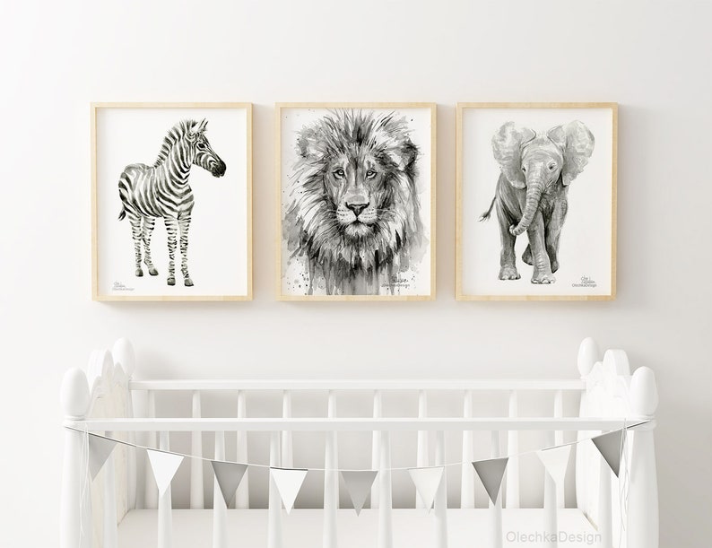 May include: Three black and white animal illustrations in wooden frames. The illustrations are of a zebra, a lion, and an elephant. The frames are hanging on a white wall above a white crib with a gray and white bunting.