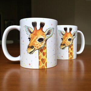 May include: Two white ceramic mugs with a colorful splatter design. Each mug features a hand-painted illustration of a giraffe with a heart-shaped pattern on its neck.