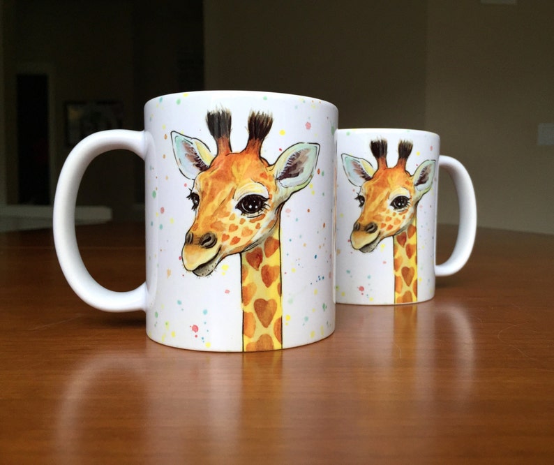 May include: Two white ceramic mugs with a colorful splatter design. Each mug features a hand-painted illustration of a giraffe with a heart-shaped pattern on its neck.